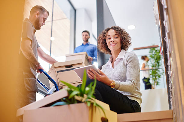 a mid adult female business owner sits on some moving boxes and looks optimistically at her new offices. Around her , co-workers and removal men are moving boxes and plants as she works on her digital tablet .
